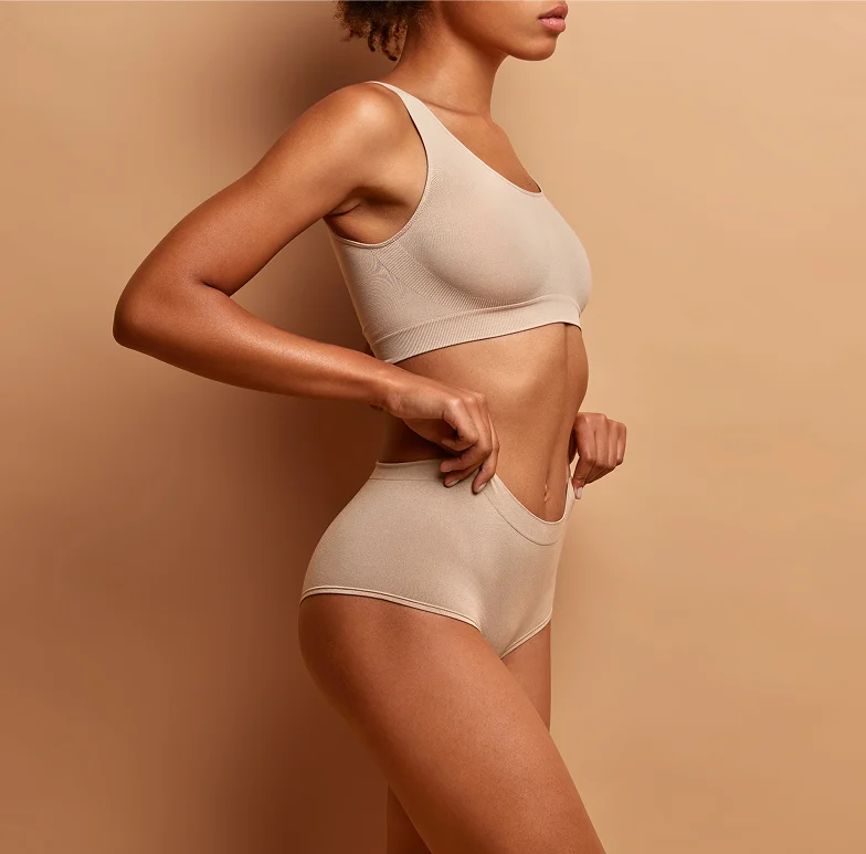 Side view of woman with curly hair wearing beige sports bra and high-waisted underwear, hand on hip against beige background.