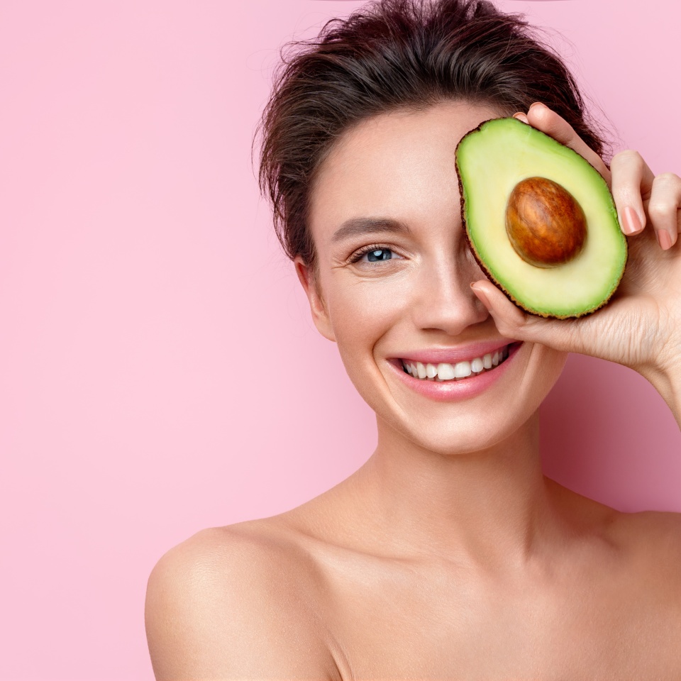 Pretty woman holds half an avocado in front of her face. Photo of attractive woman with perfect makeup on pink background. Beauty & Skin care concept