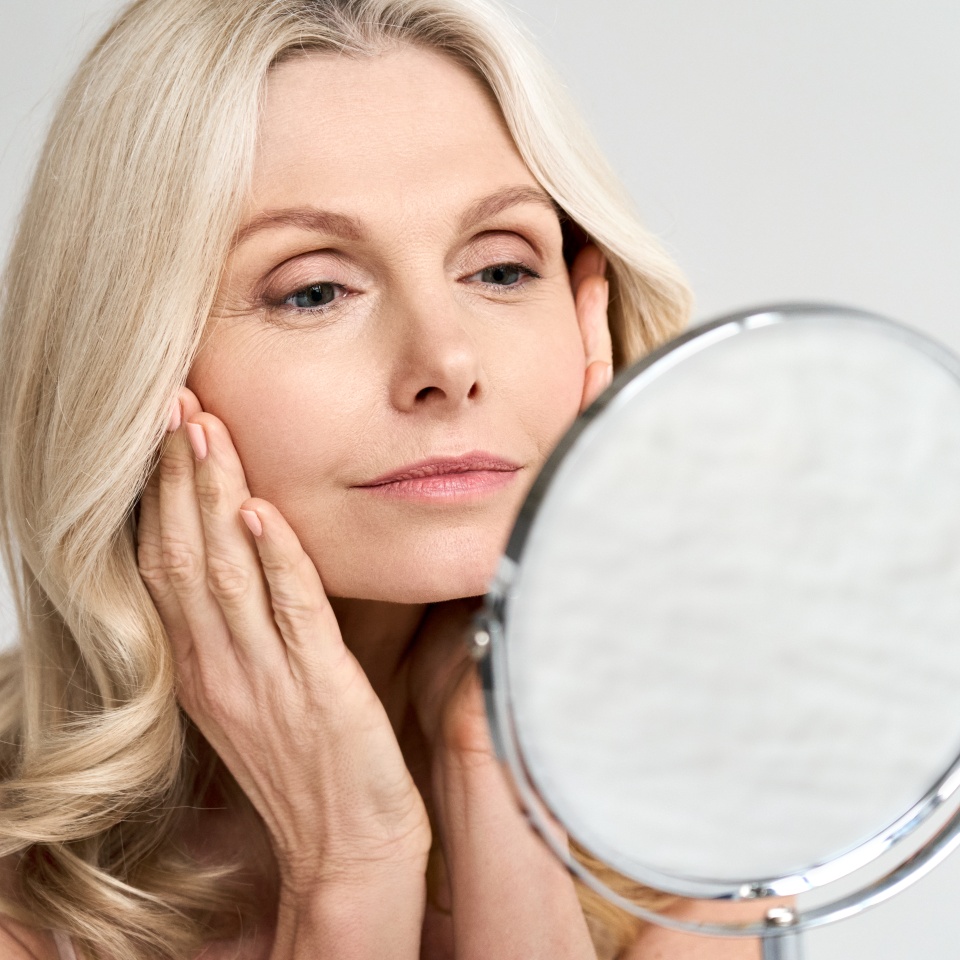Closeup portrait of gorgeous happy middle age woman looking at mirror touching her skin enjoying treatment for dry skin. Advertising of antiaging beauty skin care products.