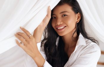 Happy Woman With Beautiful Face Smiling, Lying On Bed. Portrait Of Beautiful Young Female With Healthy Hair, Fresh Soft Facial Skin And Perfect White Smile On White Bedding. High Resolution.